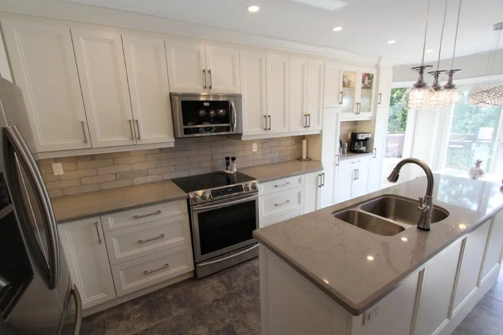 Modern kitchen with white cabinetry, gray countertops, stainless steel appliances including a stove with an overhead microwave, and a large island featuring a double sink and a curved faucet, illuminated by hanging pendant lights.