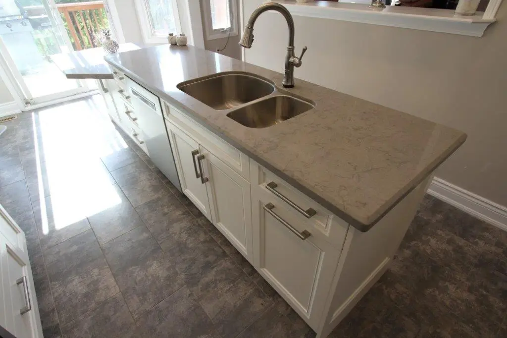 Modern kitchen island with a grey marble countertop, stainless steel double sink, brushed metal faucet, white cabinetry, and a grey tiled floor with sunlight coming through a glass door in the background