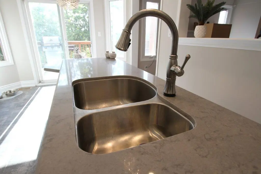 Close-up of a double stainless steel kitchen sink with a curved faucet installed in a polished stone countertop, natural light coming from large windows in the background.