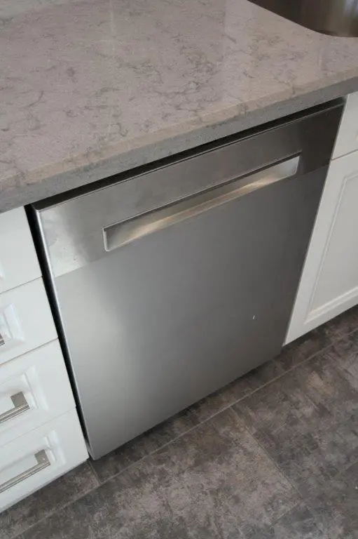 Stainless steel built-in dishwasher under a light gray marble countertop, surrounded by white cabinetry with silver handles, on a dark textured tile floor.