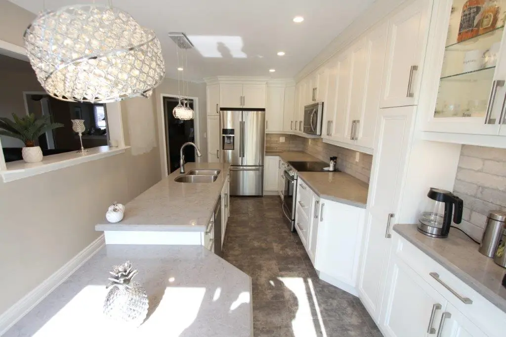 Bright modern kitchen with white cabinets, gray countertops, stainless steel fridge and appliances, two sinks on the island, decorative pendant lights, and a tiled backsplash.