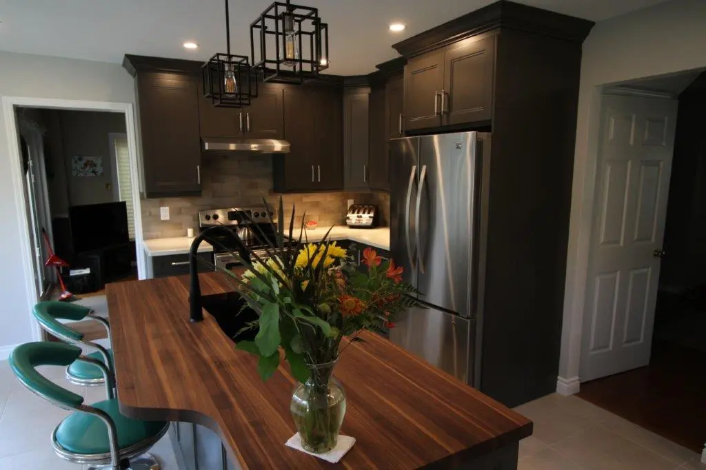 Modern kitchen with dark cabinets, stainless steel refrigerator and stove, a wooden island countertop with a vase of yellow and orange flowers, and green cushioned bar stools.