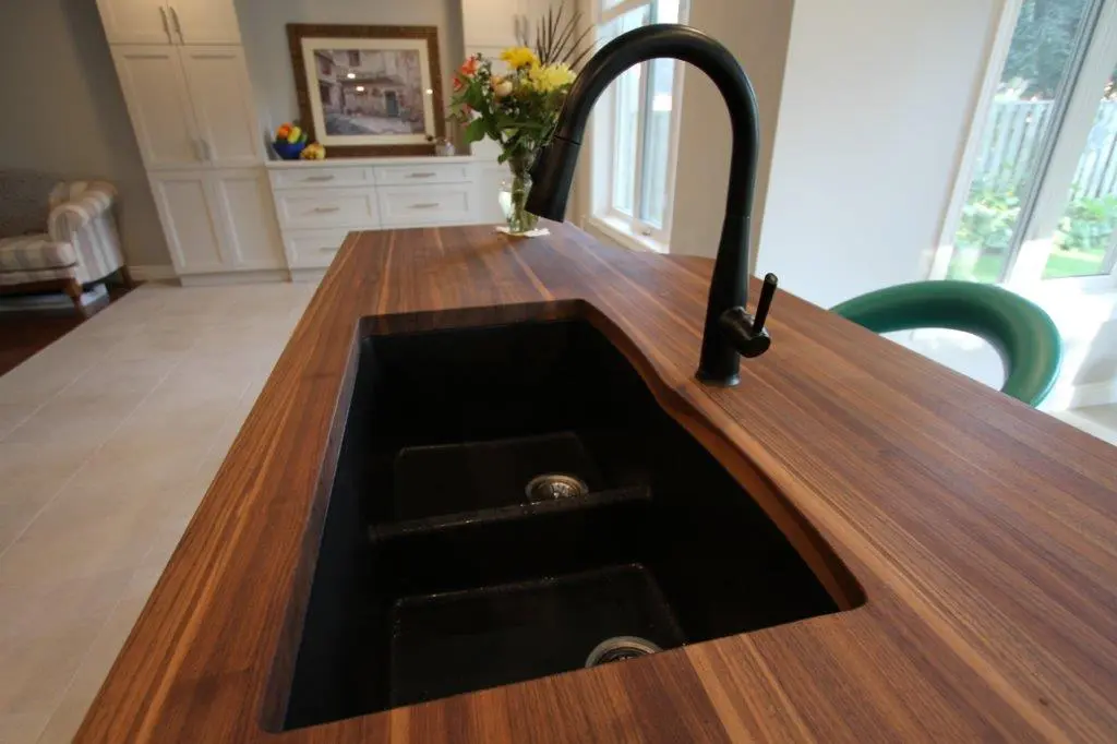 Close-up of a modern black double kitchen sink with a curved black faucet set in a wooden countertop, with a vase of flowers, white cabinetry, a framed picture, and green chair visible in the background near large windows.