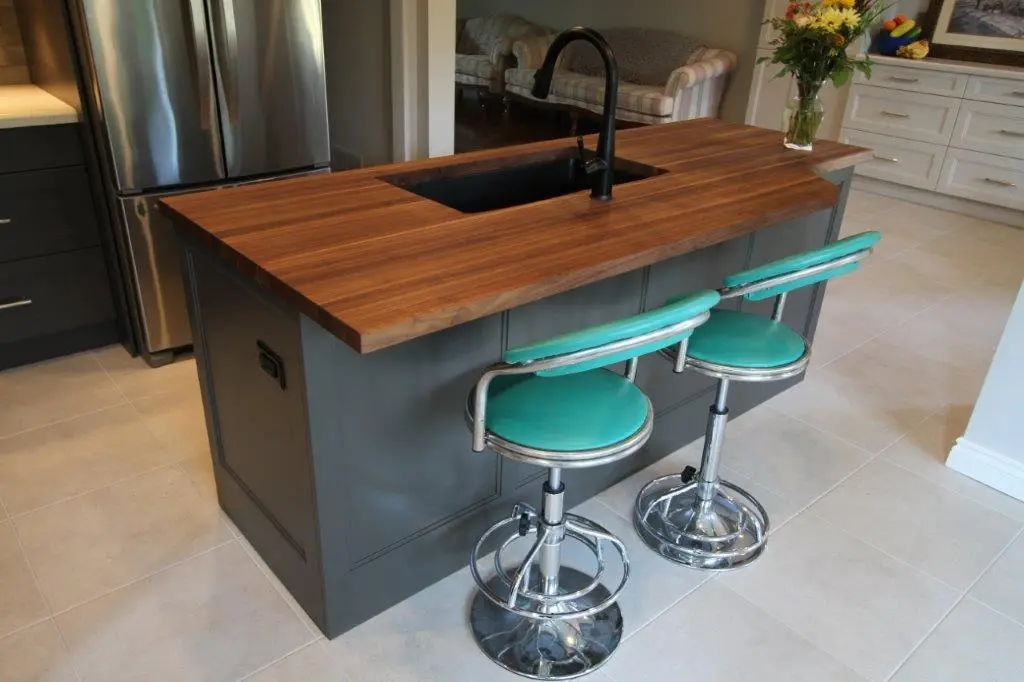 Modern kitchen island with wood countertop and black sink faucet, accompanied by two turquoise bar stools with chrome bases, set on light-colored tiled floor near a stainless steel refrigerator and a living room in the background