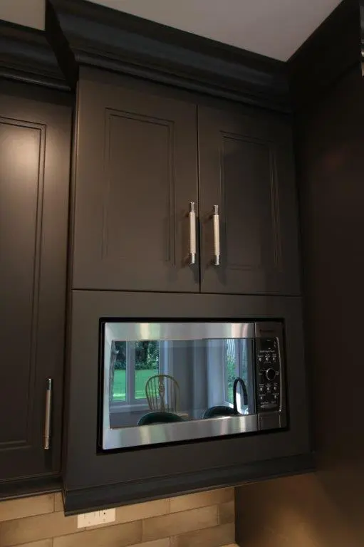 Dark kitchen cabinets with silver handles above and beside a built-in microwave reflecting a window, chair, and some greenery outside.