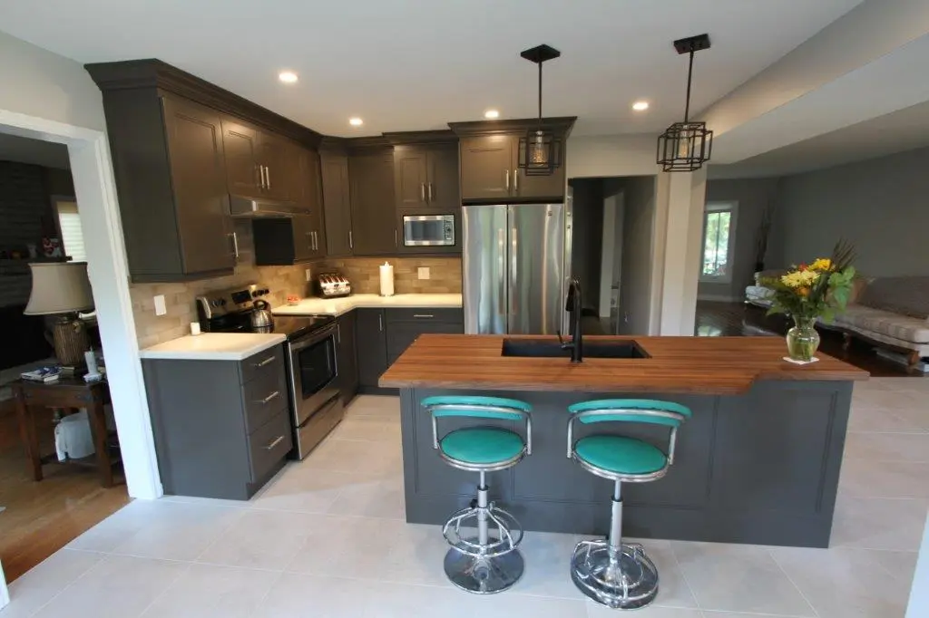 Modern kitchen with dark gray cabinets, stainless steel appliances, a wooden island countertop with a black sink, two turquoise bar stools, and pendant lights above the island; adjacent to a living area with a sofa and window.