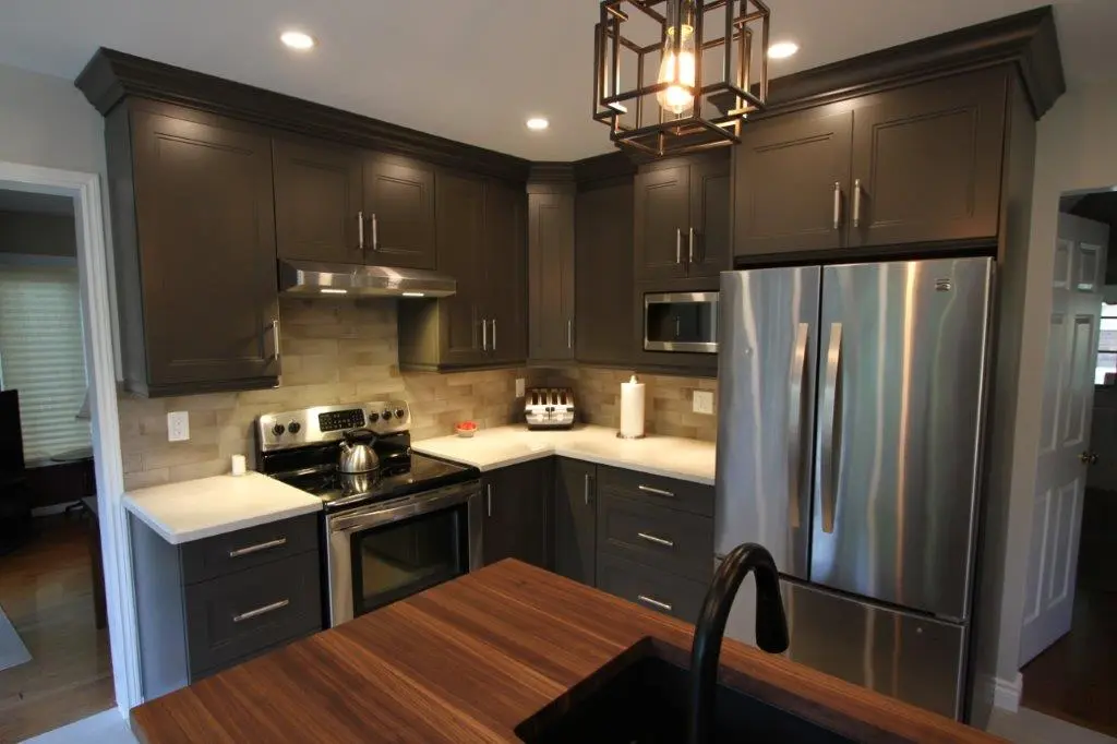 Modern kitchen with dark gray cabinets, stainless steel appliances including a refrigerator and oven, light-colored countertop, wooden island with a black faucet, and overhead recessed lighting.