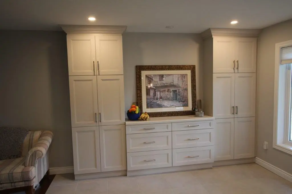 White built-in cabinets with multiple drawers and doors centered on a beige wall under recessed lighting, topped with a framed painting, a bowl of fruit, and a bunch of bananas, with a striped armchair partially visible on the left and a window on the right.