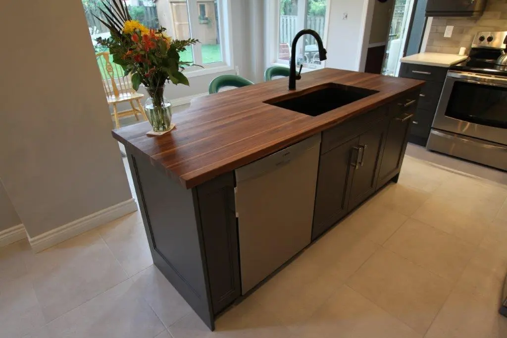 Modern kitchen island with a dark wood countertop, black sink and faucet, dishwasher, and dark cabinetry, with a vase of colorful flowers on the corner and light-colored tile flooring.