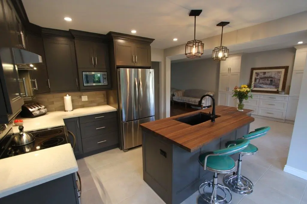Modern kitchen with dark cabinets, stainless steel refrigerator, wooden island with black sink and faucet, two turquoise bar stools, and pendant lights, adjacent to a room with a sofa and white cabinetry with a framed painting and a vase of flowers.