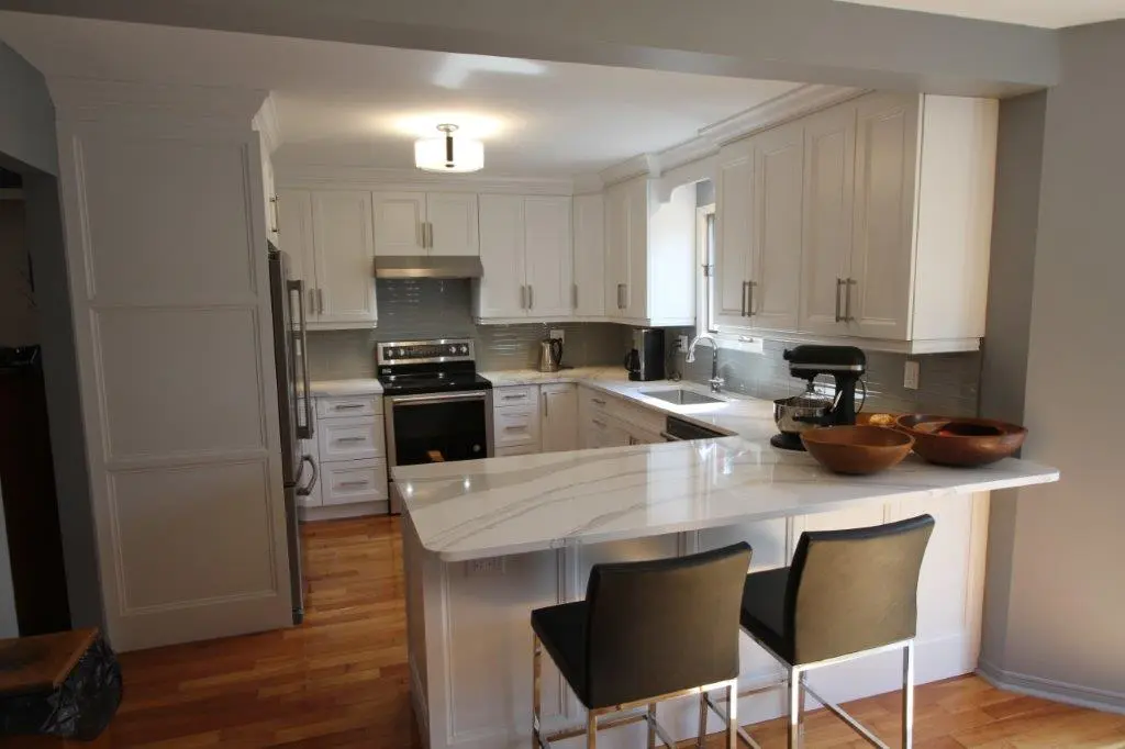 Modern kitchen with white cabinetry, stainless steel appliances, a white marble countertop island with two dark counter stools, wooden bowls on the island, and hardwood flooring