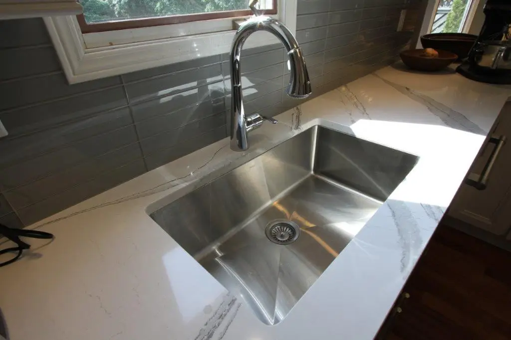 Modern kitchen sink with a stainless steel basin and a sleek chrome faucet set into a white marble countertop with gray veining, with a window and gray subway tile backsplash in the background.