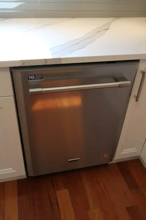 Stainless steel built-in dishwasher with a horizontal handle installed under a white marble countertop with gray veining, surrounded by white cabinets and resting on a wooden floor.