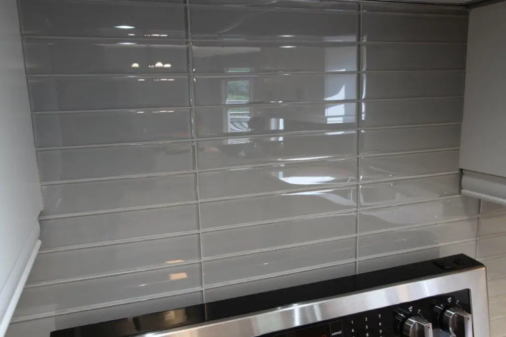 Glossy light gray rectangular subway tiles arranged in horizontal rows on a kitchen backsplash above a stainless steel stove with control knobs.