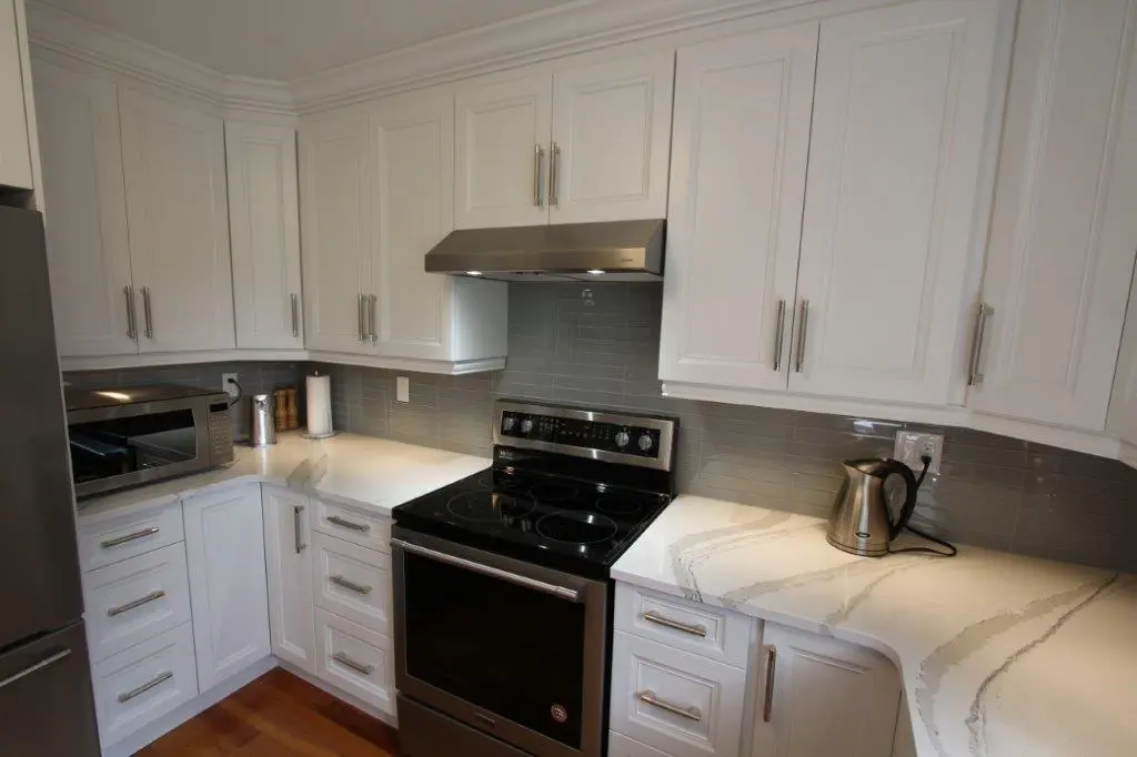 Modern kitchen corner with white cabinets, stainless steel electric stove and hood, microwave on the left countertop, and a stainless steel electric kettle on the right countertop with gray subway tile backsplash and wooden floor.