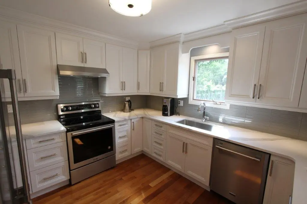 Modern kitchen with white cabinets, stainless steel appliances including an oven and dishwasher, a gray tiled backsplash, white countertops, a stainless steel sink under a window, and wooden flooring.