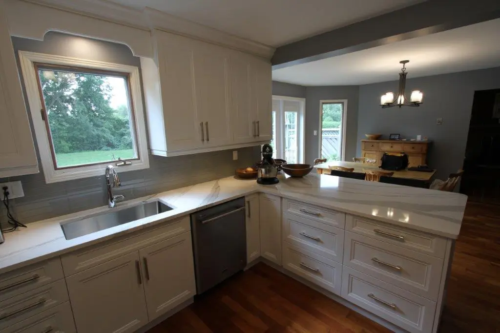Modern kitchen with white cabinets, gray backsplash, marble countertops, stainless steel sink and dishwasher, wooden floor, and a dining area with wooden table, chairs, and a chandelier in the background