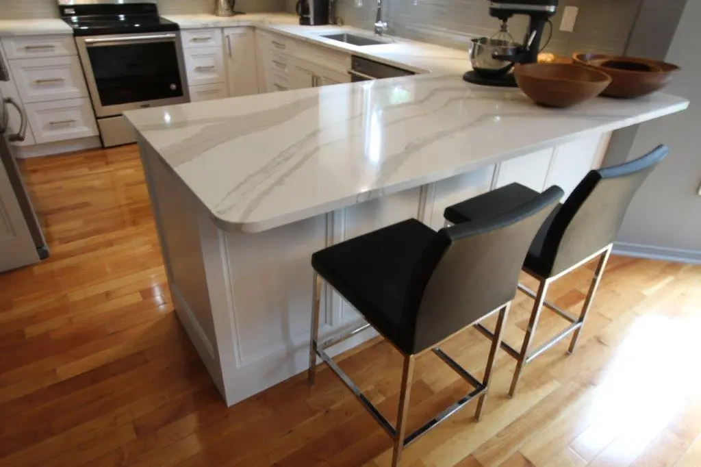 Kitchen interior featuring a white marble countertop island with two modern black chairs with chrome legs, hardwood floor, white cabinets, and kitchen appliances including an oven and a mixer on the counter.
