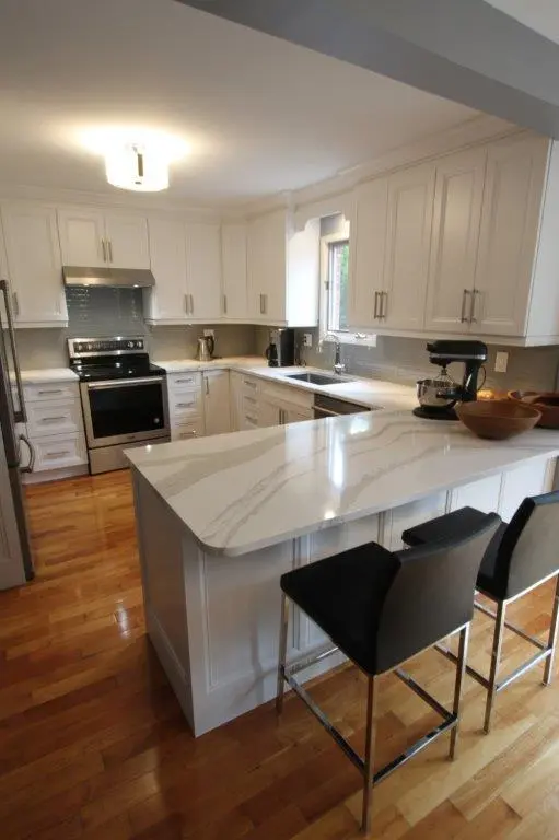 Modern kitchen with white cabinets, stainless steel appliances, a marble countertop island with two black bar stools, and wooden flooring.