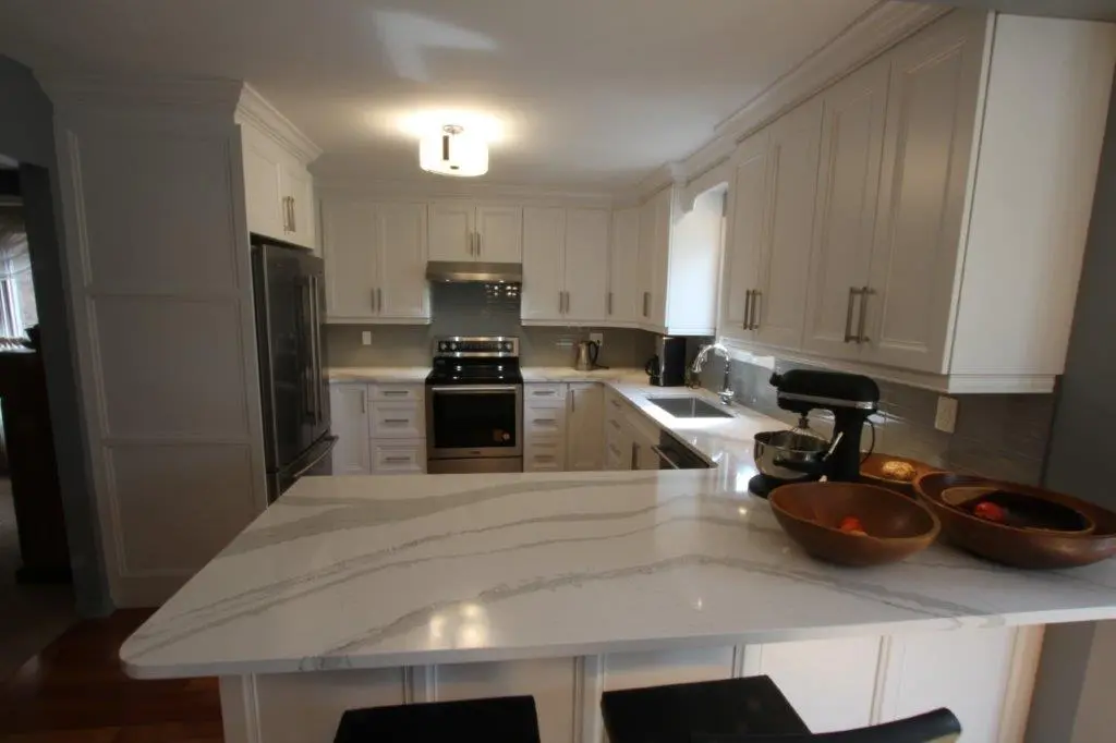 Modern white kitchen with marble countertop island, stainless steel appliances including oven and refrigerator, white cabinetry, and wooden fruit bowls on the counter