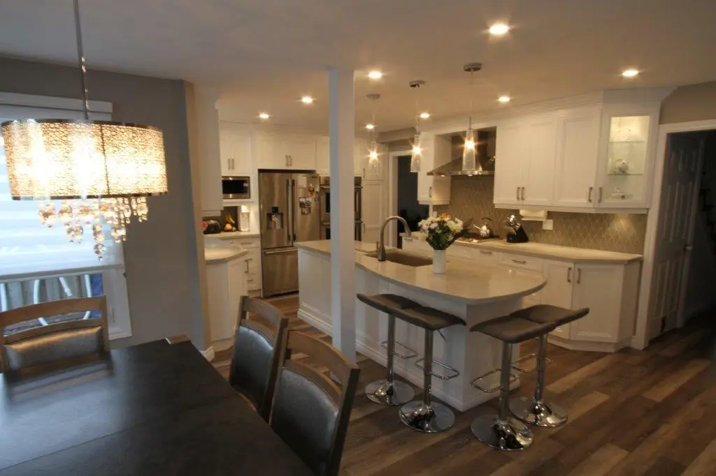 Modern kitchen and dining area with white cabinetry, stainless steel appliances, a lighted island with three bar stools, hanging pendant lights, and a chandelier over a dark dining table with chairs on wood flooring.