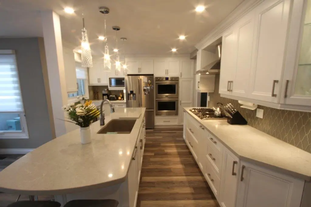 Modern kitchen with white cabinets, a large central island with a sink, stainless steel refrigerator and double ovens, gas cooktop, pendant lights above the island, and wooden floor.