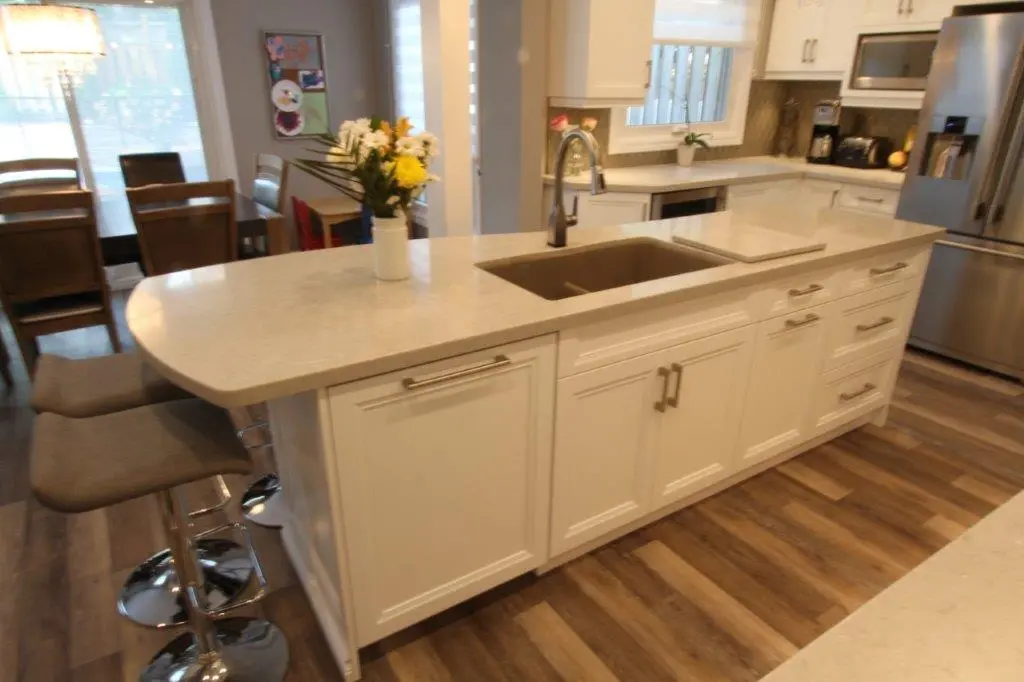 Modern kitchen interior featuring a white island with a built-in sink, three gray bar stools, a vase with yellow and white flowers on the island countertop, white cabinetry, stainless steel refrigerator, microwave, and coffee maker, with wooden flooring and a dining table in the background.