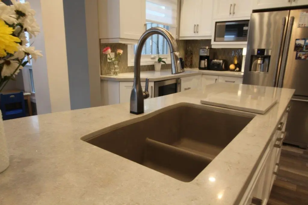 Modern kitchen with a large beige countertop featuring a built-in double sink and a high-arc stainless steel faucet, white cabinets, stainless steel refrigerator, microwave, coffee maker, and flowers on the counter.