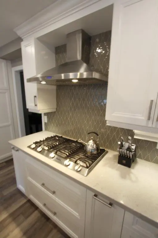 Modern kitchen countertop with a stainless steel gas stove and kettle, white cabinetry, a stainless steel range hood, and a knife block against a gray tiled backsplash.