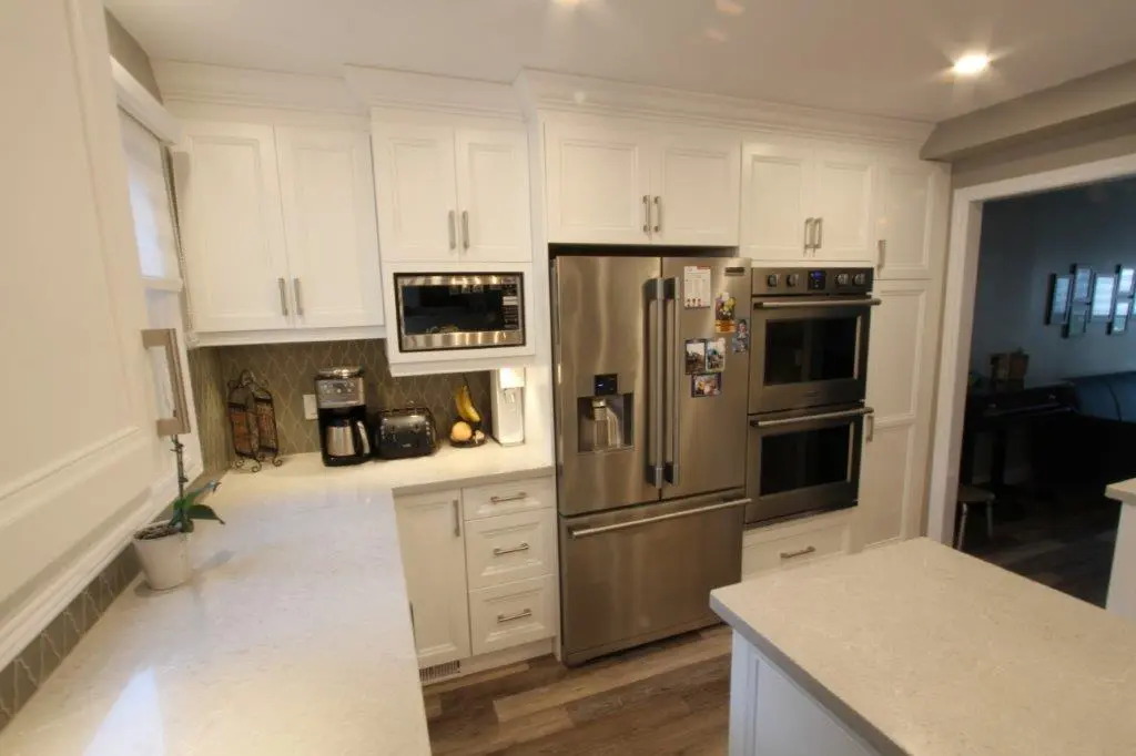 Modern kitchen interior with white cabinetry, stainless steel double ovens and refrigerator, white quartz countertops, and wood flooring, with coffee maker and toaster on the counter.