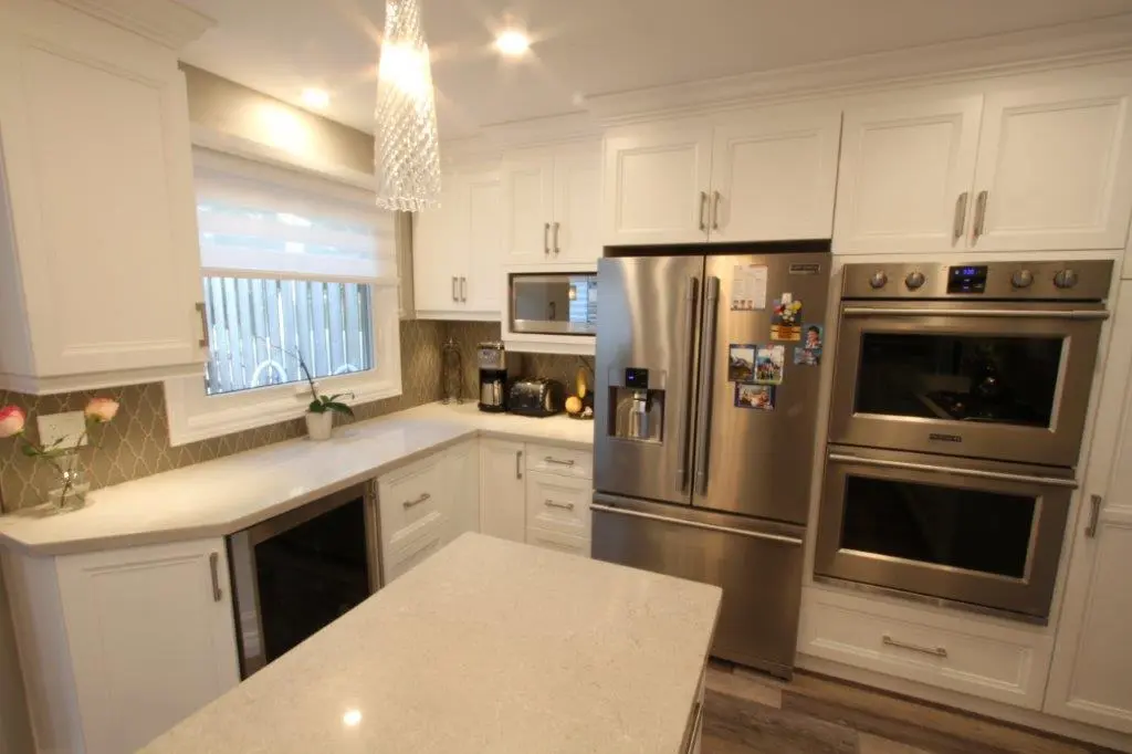 Modern kitchen with white cabinetry and quartz countertops, featuring a stainless steel refrigerator with water dispenser, double wall oven, built-in microwave, toaster, coffee maker, and a window with a roller shade, illuminated by ceiling lights and a hanging pendant light above the island.