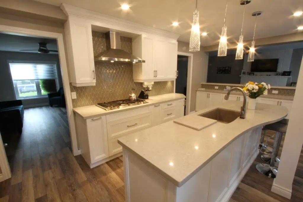Modern kitchen with white cabinets, a large light-colored countertop island with a sink and a vase of flowers, stainless steel range hood above a gas stove, pendant lights hanging from the ceiling, and wood flooring extending into an adjacent living room with a window.