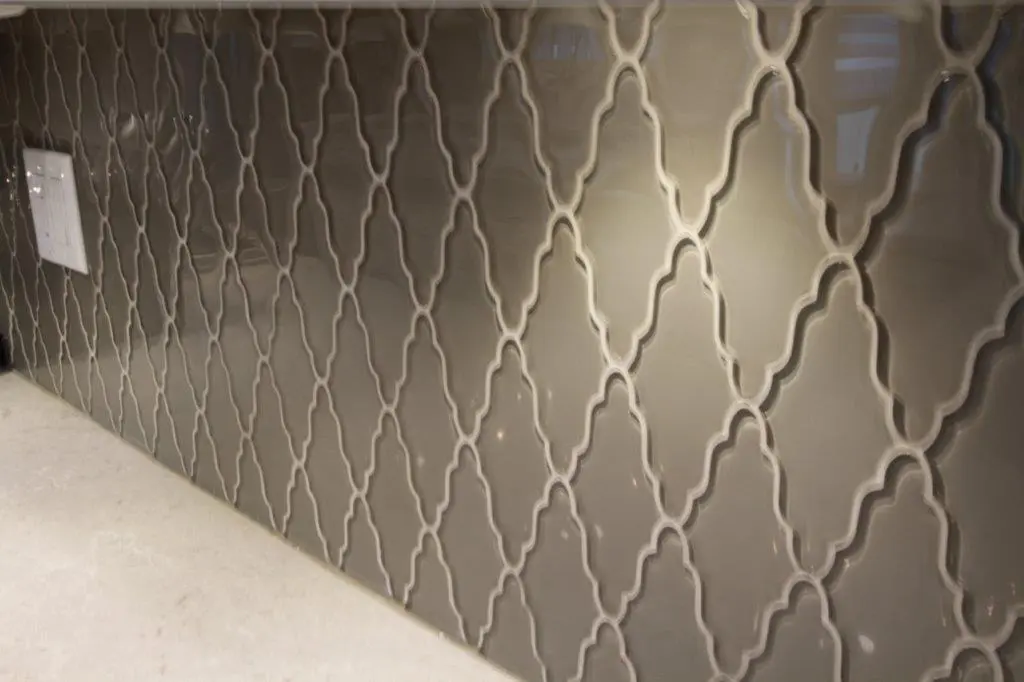 Glossy gray ceramic tiles with an intricate interlocking geometric pattern covering a wall, with a white electrical outlet on the left and a light-colored countertop below.