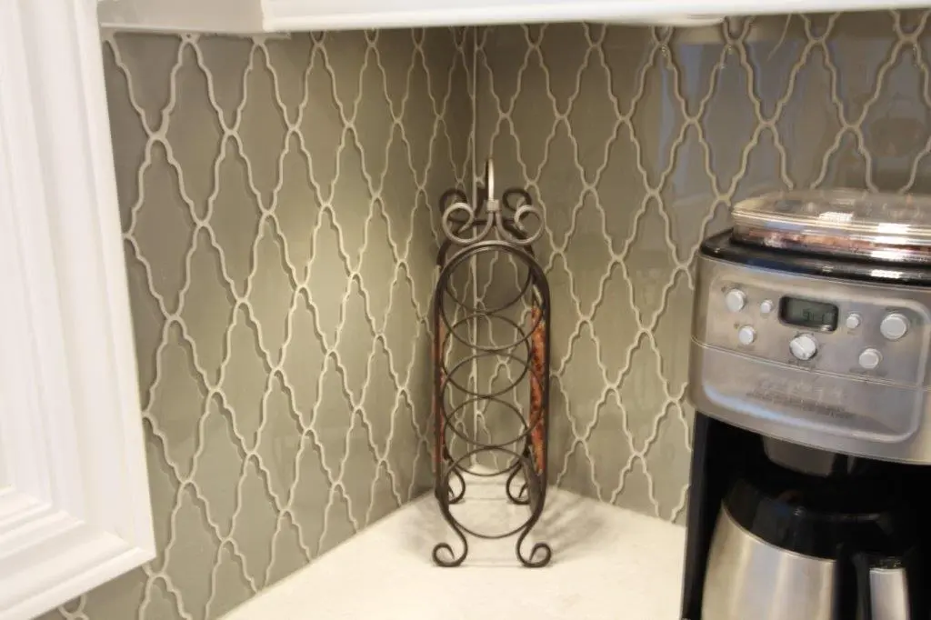 Empty decorative black metal wine rack on a kitchen counter corner next to a stainless steel coffee maker with a digital display, against a tiled backsplash with a geometric pattern.