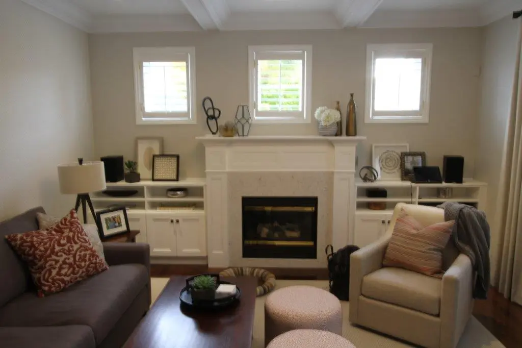 Cozy living room with a white fireplace centered between two windows, flanked by white built-in shelves holding decorative items and speakers, a gray sofa with patterned pillows on the left, a beige armchair with a striped pillow and a gray throw on the right, and a wooden coffee table with decor in the foreground.