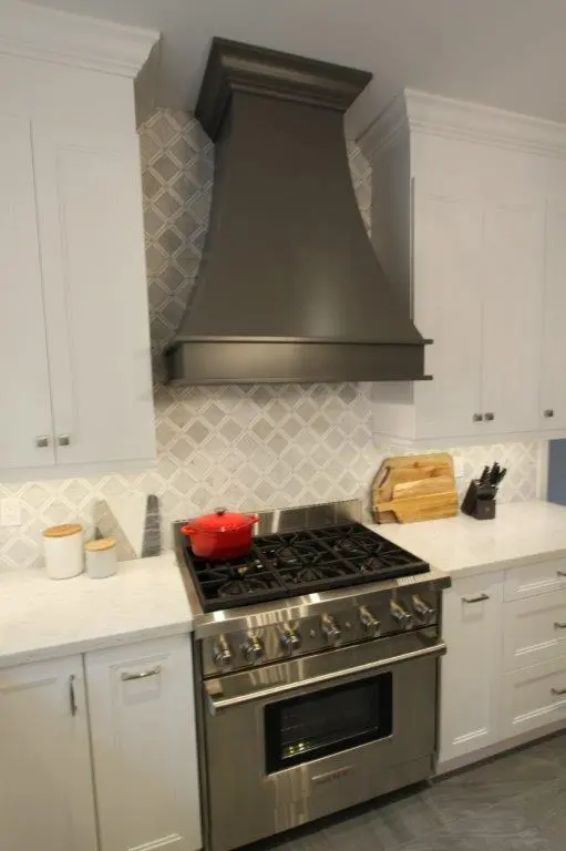 Modern kitchen stove with six burners, a stainless steel oven, a black range hood, white cabinetry, white countertop, cutting boards, a set of knives, and a red pot on the stove.