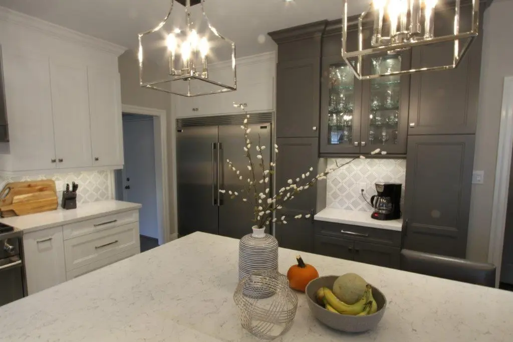Modern kitchen with white and dark gray cabinets, stainless steel refrigerator, marble countertops, two pendant lights, a striped vase with branches, a bowl of bananas and cantaloupe, and a small pumpkin on the kitchen island.