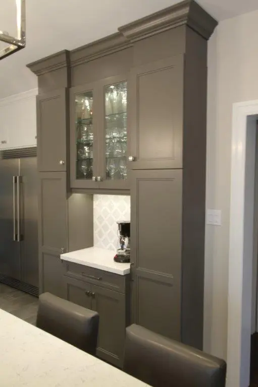 Tall gray kitchen cabinets with glass doors displaying glassware, a white countertop with a coffee maker, set against a patterned backsplash, adjacent to a stainless steel refrigerator and dark leather chairs in the foreground.