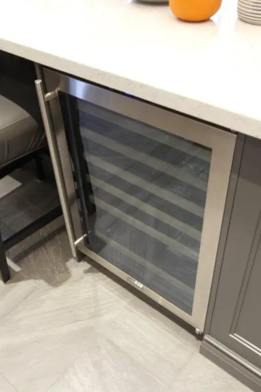 Built-in stainless steel wine cooler with glass door beneath a white countertop in a modern kitchen.