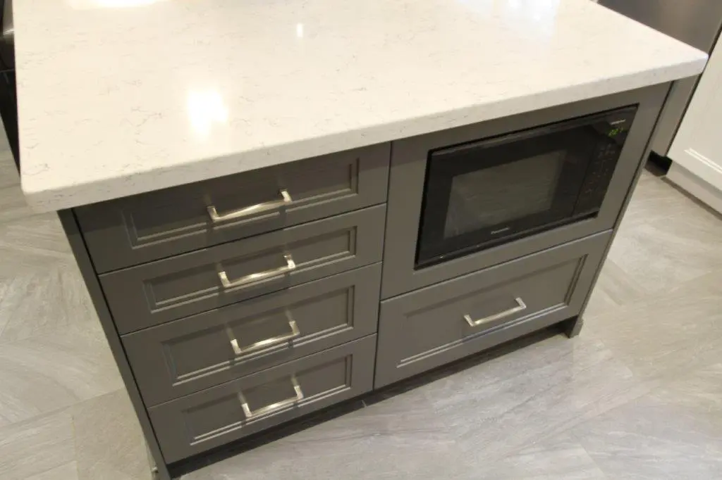Gray kitchen island with four drawers on the left and a built-in black microwave with a drawer beneath it on the right, topped with a white marble countertop, situated on a light gray tiled floor.