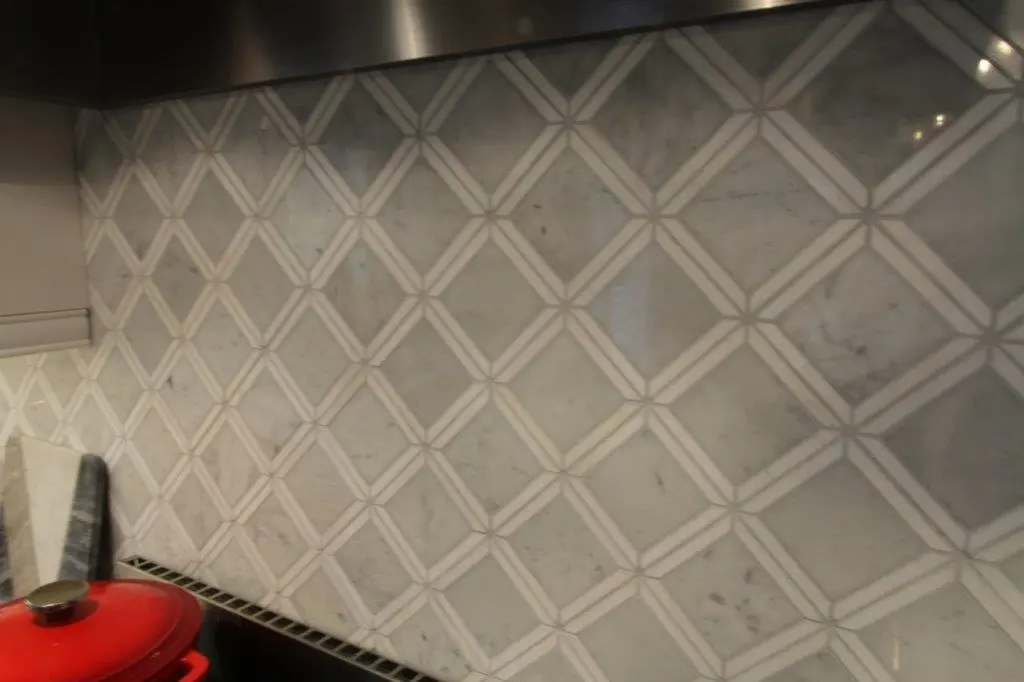 Kitchen backsplash with a diamond-patterned tile design in light gray and white tones, and a red pot on the stovetop in the lower left corner.