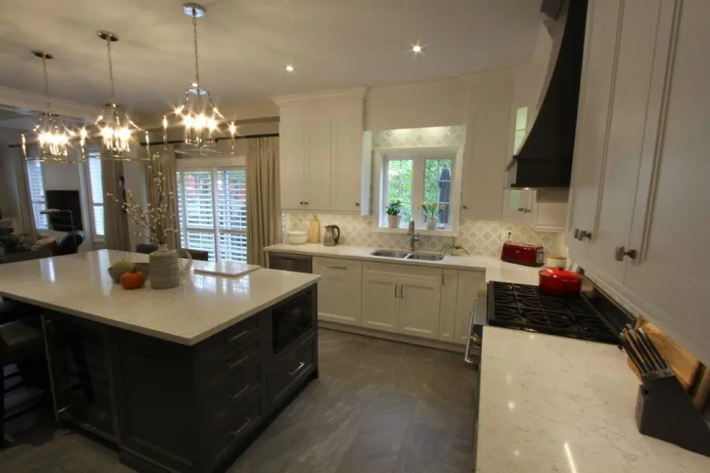 Modern kitchen with a large central island featuring a white countertop, dark cabinetry, pendant lighting, white cabinets along the walls, a window above the sink, and stainless steel appliances including a stove with a red pot and a red toaster on the counter.