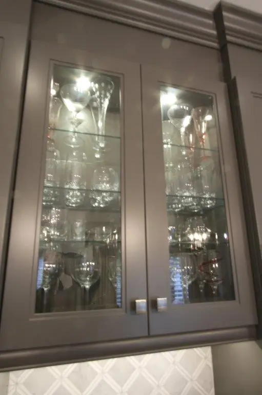 Glass-fronted kitchen cabinet filled with various glassware including wine glasses and champagne flutes, with reflections from a chandelier visible on the glass doors.