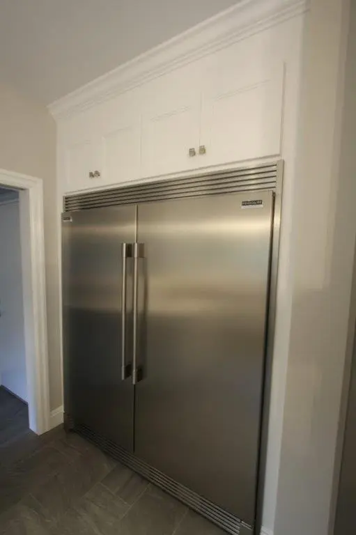 Large built-in stainless steel refrigerator with double doors, integrated into white cabinetry in a kitchen with gray tiled floor.