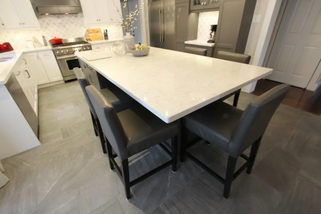 Modern kitchen with a white marble island countertop surrounded by black leather bar stools, stainless steel stove and appliances, and white cabinets.