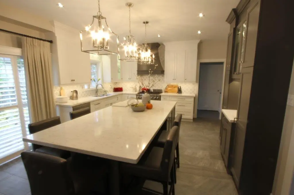 Modern kitchen with a large white marble island surrounded by black chairs, white cabinetry, hanging pendant lights, and stainless steel appliances.