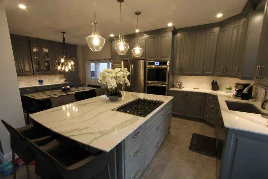Modern kitchen with gray cabinetry, a white marble island featuring a built-in gas cooktop, three pendant lights hanging above the island, a dining area with a contemporary chandelier, stainless steel double ovens and refrigerator, and a sink area with a coffee maker and small plants.