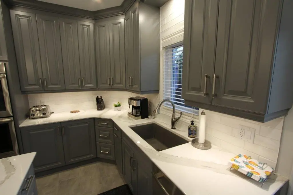 Modern kitchen corner with gray cabinets, white marble countertops, a stainless steel sink under a window with blinds, a toaster, coffee maker, knife block, paper towel holder, and a colorful towel with pineapple print on the counter.