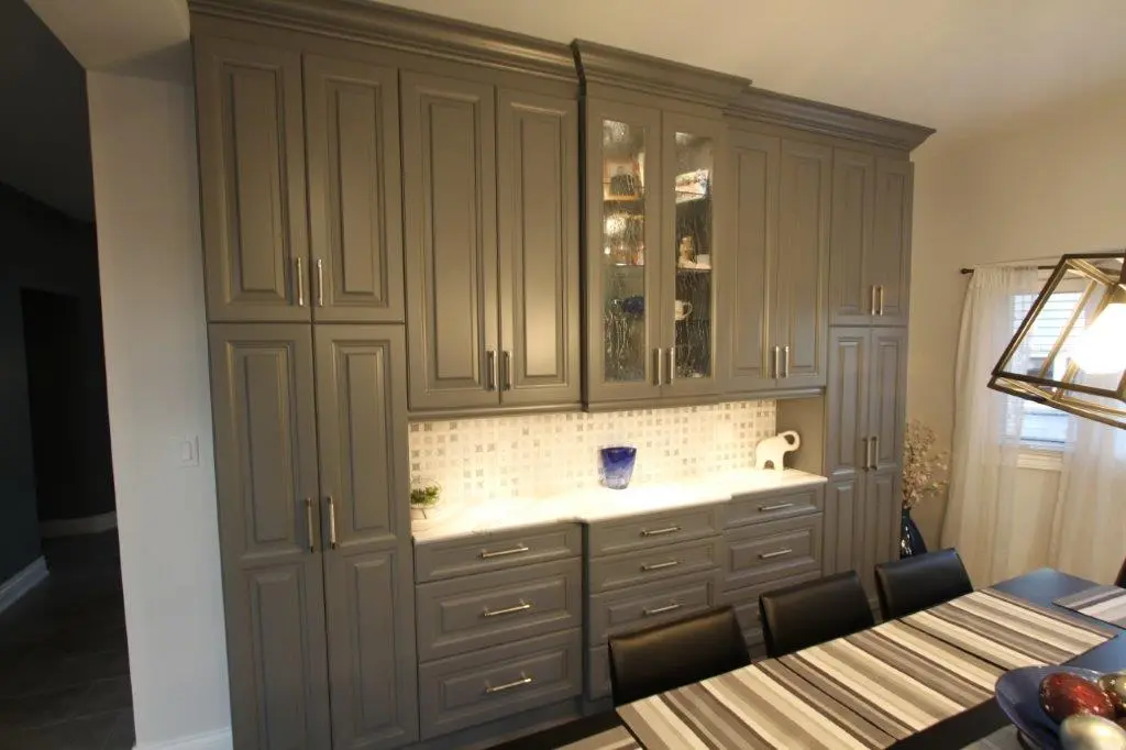 A dining room with gray paneled cabinets featuring glass-paneled upper doors and silver handles, a white marble countertop with decorative items, a striped tablecloth on the dining table, black leather chairs, and a large modern pendant light, with a window and light curtains in the background.