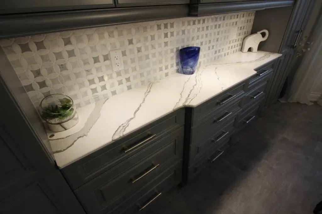 Marble countertop with gray veining set against white patterned tile backsplash, featuring a glass bowl with succulents, a blue glass vase, and a white ceramic elephant figure on gray kitchen cabinets with silver handles.
